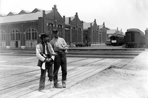 Dwóch żołnierzy stoi obok budynku Pullman i trenuje wozy z zamkniętymi ramionami i butelką alkoholu podczas strajku w Chicago w 1894 roku